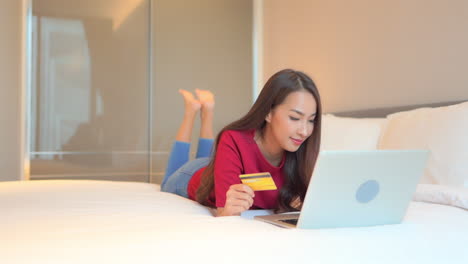 a young asian woman shopping online using credit card and laptop computer in bed of family home