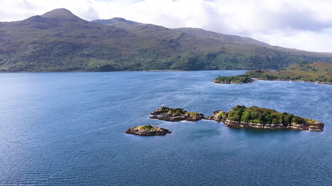 Drone glides above blue sea toward lush green islands, distant mountains, and partly cloudy sky