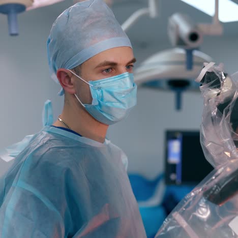 young male surgeon near medical equipment. side view portrait of a doctor in blue uniform looking into modern microscope in the operating room.