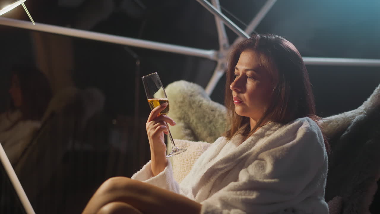 Depressed young woman drinks alcoholic drink in glass sitting in armchair at night in hotel room. Unpleasant feeling of loneliness and heartbreaking pain slow motion
