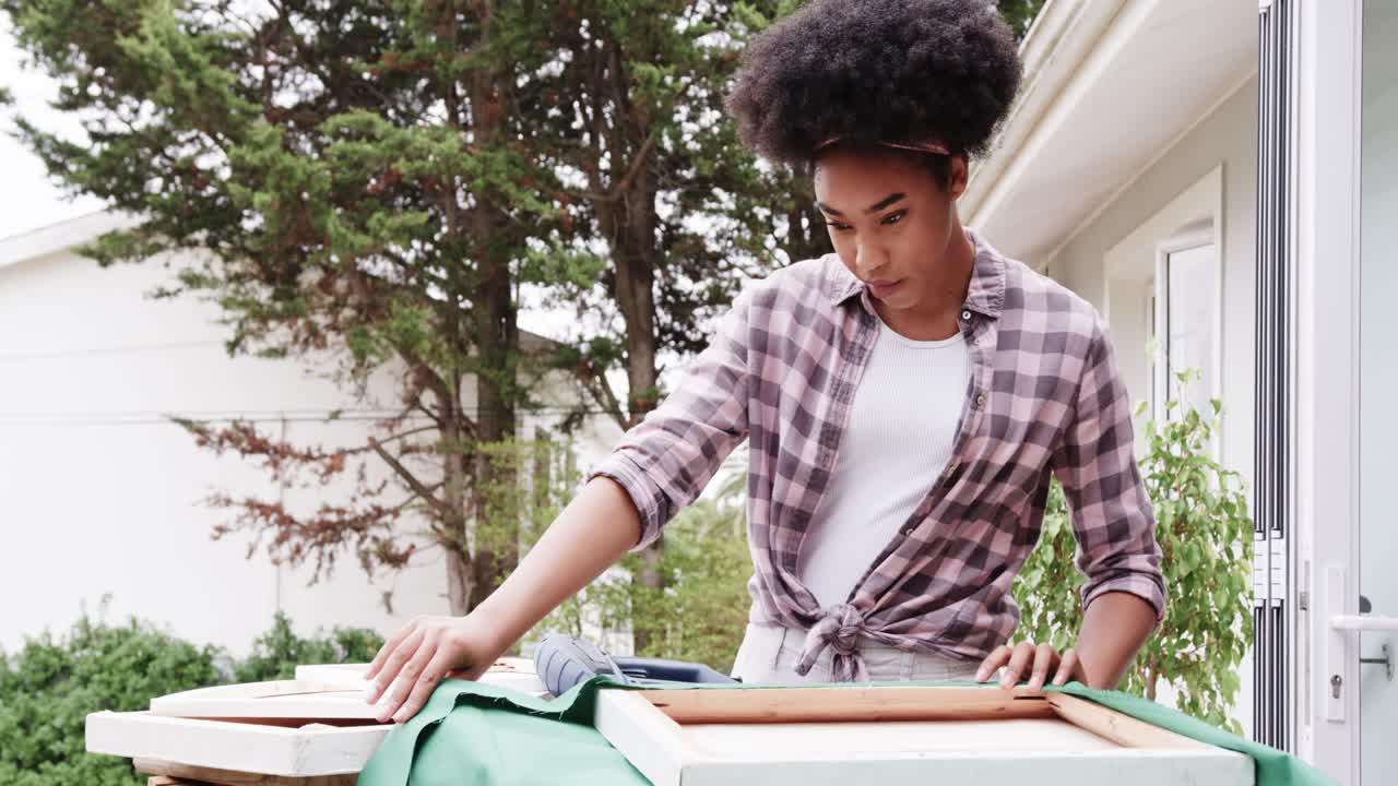 African American woman stretching green fabric on wooden frames at backyard table, stapler visible