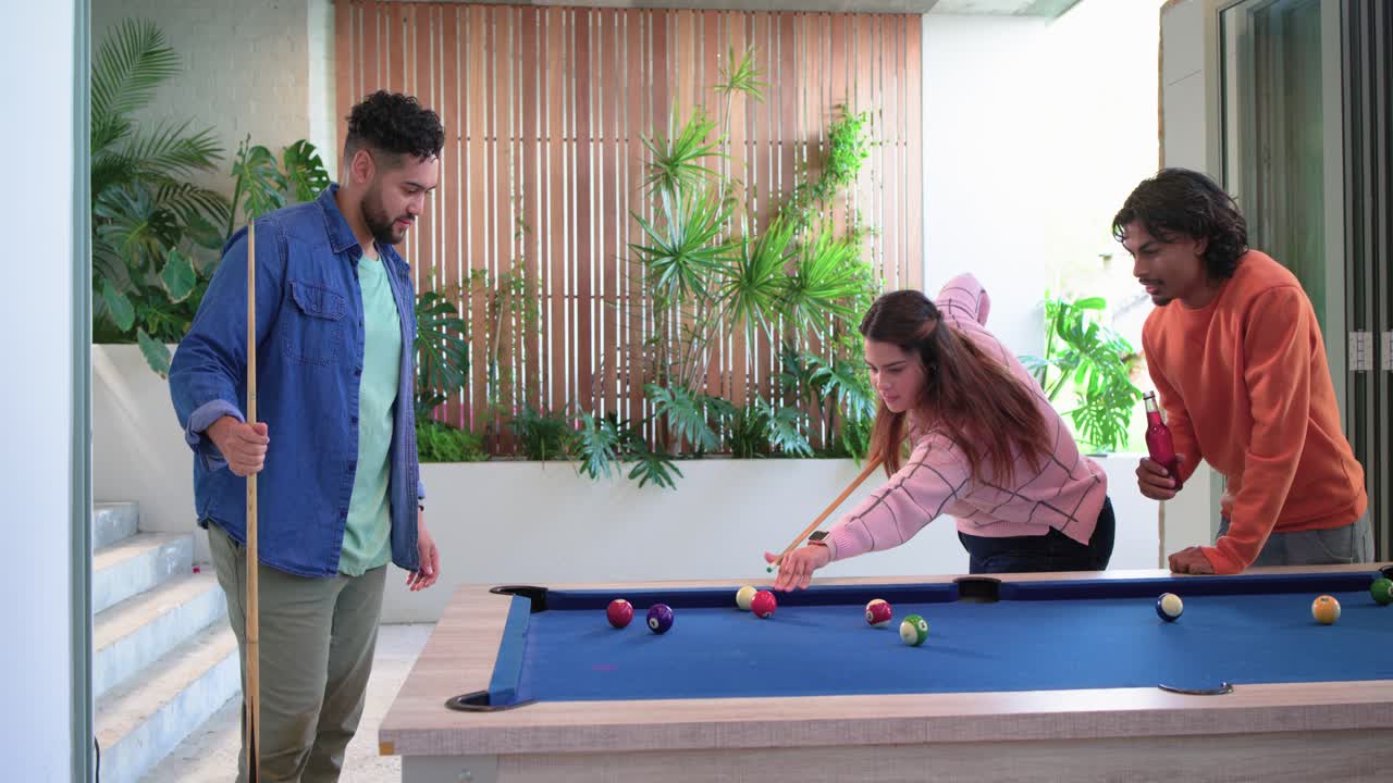 Diverse friends joking about cues at blue-felt pool table swapping tips by beer bottle, lining shot