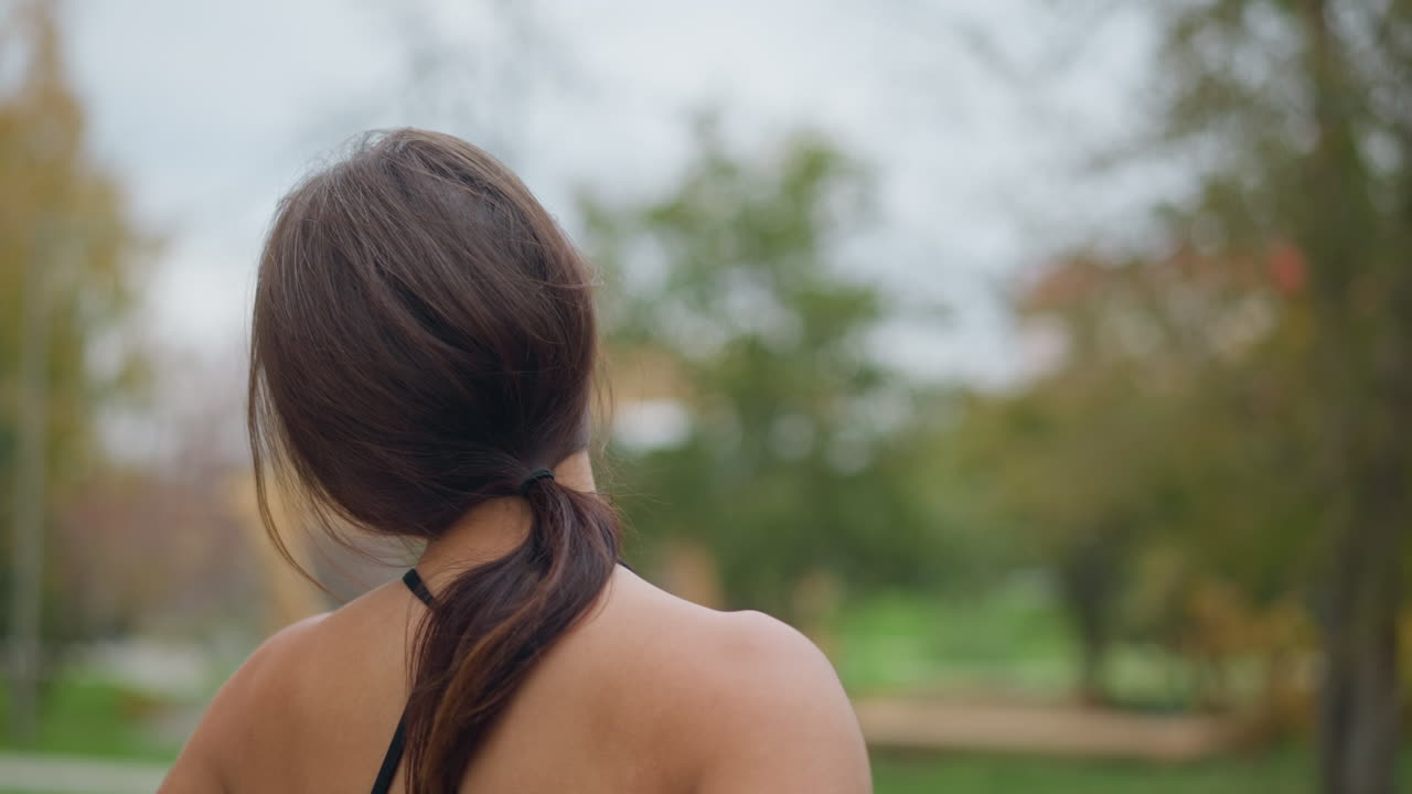Back view of woman stretching neck gently in park with trees in blurred background, capturing peaceful outdoor stretching and neck flexibility in a natural setting
