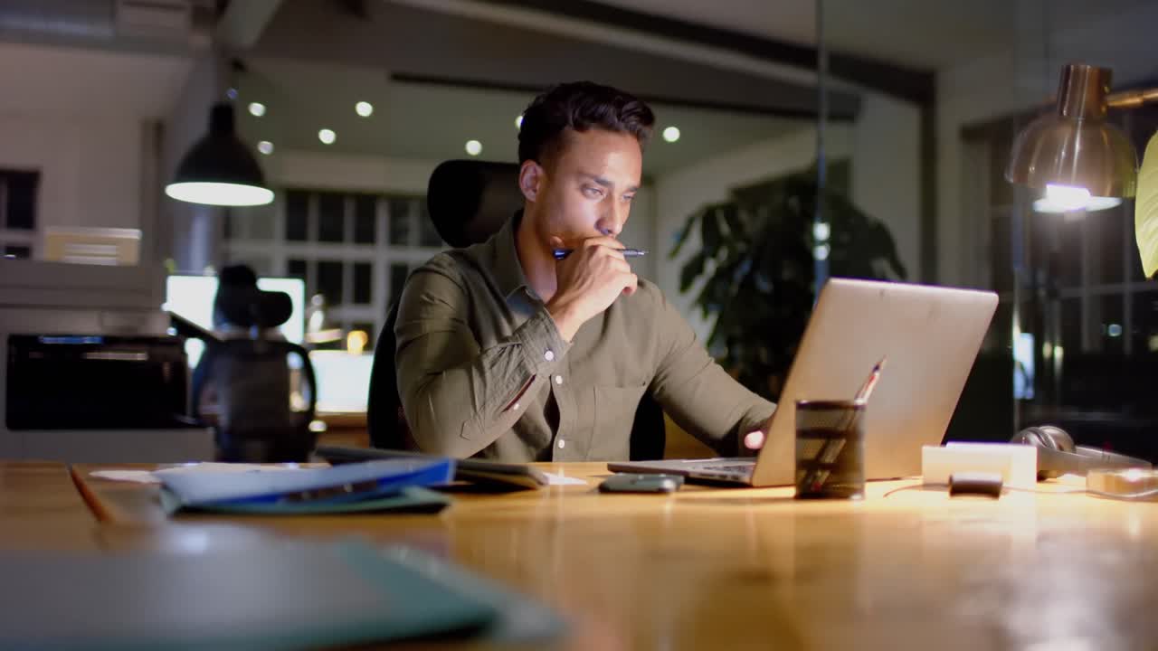 Focused biracial businessman at desk using laptop in office at night, slow motion