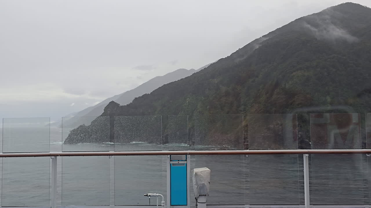 Ein Kreuzfahrtschiff im Milford Sound navigiert auf ruhigem Wasser unterhalb von nebelverhangenen, hoch aufragenden bewaldeten Klippen des Fiordland an einem trüben, friedlichen Tag in Neuseeland