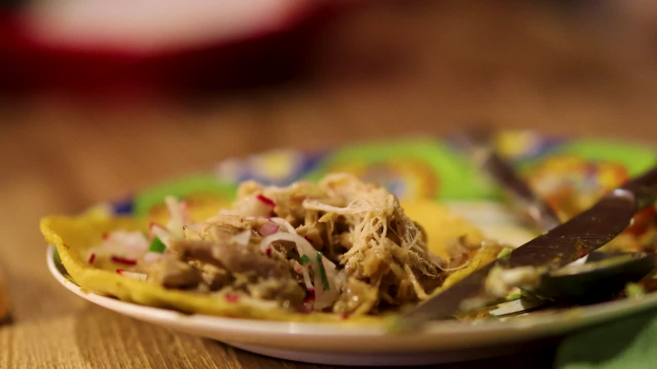 Et nærbillede af en gaffel, der løfter strimlet pulled pork fra en taco på en farverig tallerken, under varm indendørs belysning, med lav dybdeskarphed.