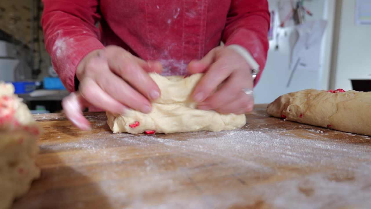 Close up of a baker kneading and shaping pastry dough mixed with pink pralines on a floured wooden table inside a traditional bakery