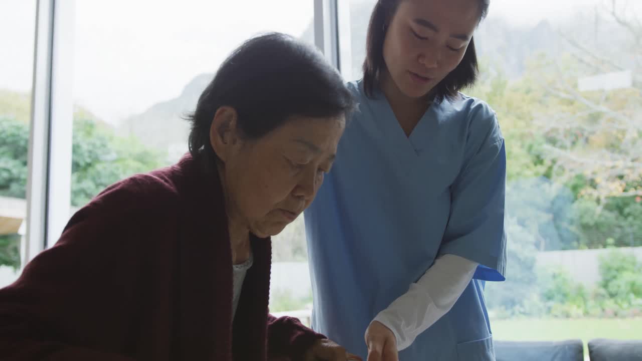 미소 짓는 아시아 여성 의사가 고령 여성 환자가 보행 프레임을 사용하여 걸을 수 있도록 돕고 있습니다.