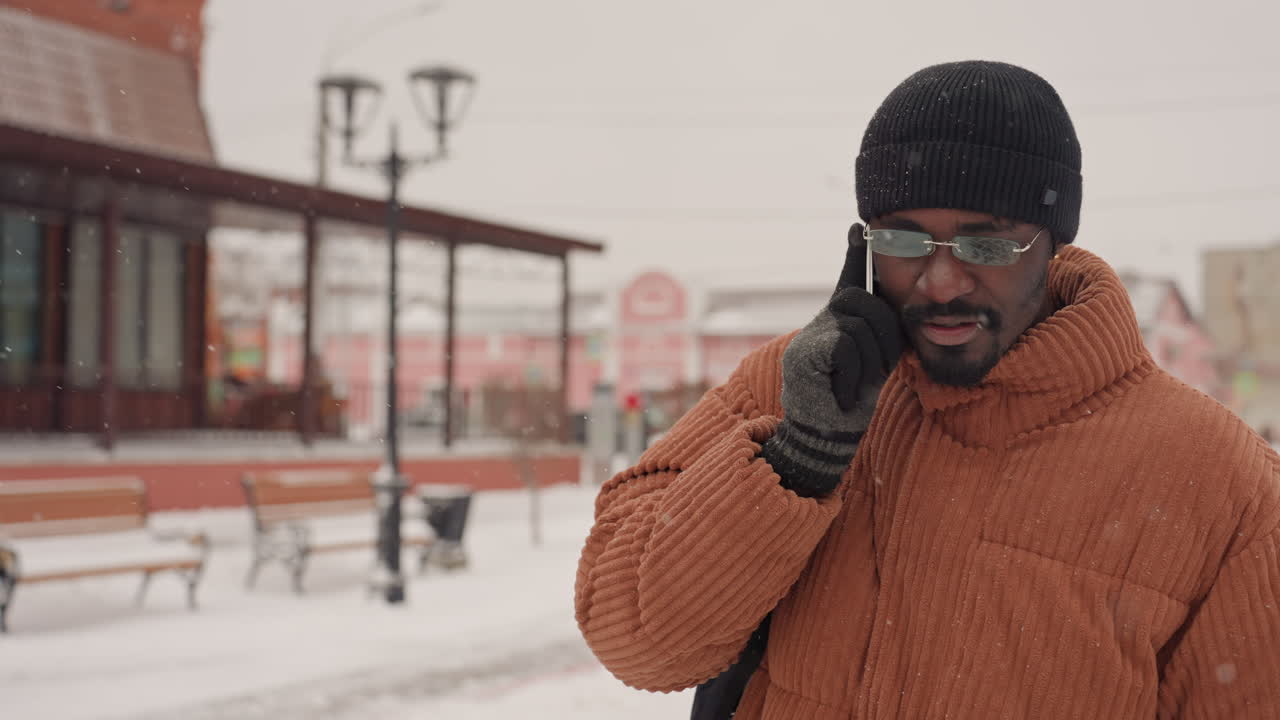 Black man walking in snow, carrying backpack, wearing orange coat and knit hat, talking on phone in urban plaza, light snowfall, thoughtful expression, commuter character, candid street lifestyle