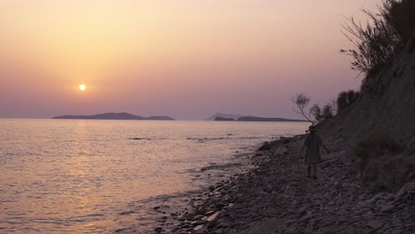 Slowmotion-shot-of-a-woman-walking-raising-her-arms-in-joy-enjoying-the-beach-at-sunset-in-Arillas,-Corfu,-Greece