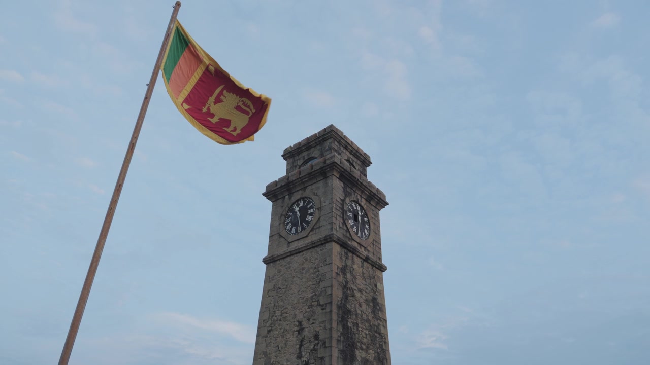 Ripresa al rallentatore da angolazione bassa della bandiera dello Sri Lanka che sventola accanto alla Torre dell'Orologio del Forte di Galle, mostrando l'architettura storica in pietra contro un cielo azzurro pallido.