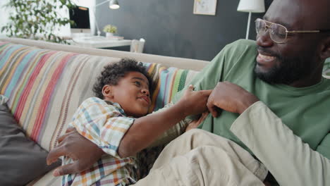 joyous african american man tickling little son on sofa at home