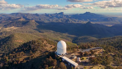 drone footage circles a large white observatory atop a forested mountain ridge, revealing sweeping views of rugged terrain under soft afternoon sunlight