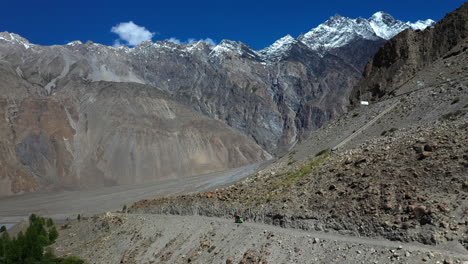 Cinematic-drone-shot-of-a-tuk-tuk-on-a-gravel-path-on-the-Karakoram-Highway-Pakistan-with-the-passu-cones-in-the-distance,-revealing-aerial-shot