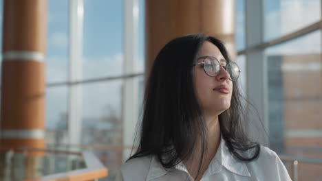 Close up of young woman in white shirt wearing eyeglasses walking indoors and looking around with thoughtful expression, decorative pillar and glass windows in blurred background