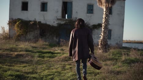 man walking with guitar in front of an abandoned house