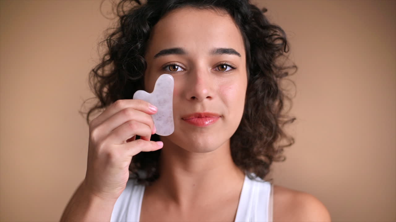 A young caucasian smiling woman is doing a facial massage using Gua Sha, looking into the camera. Slow motion