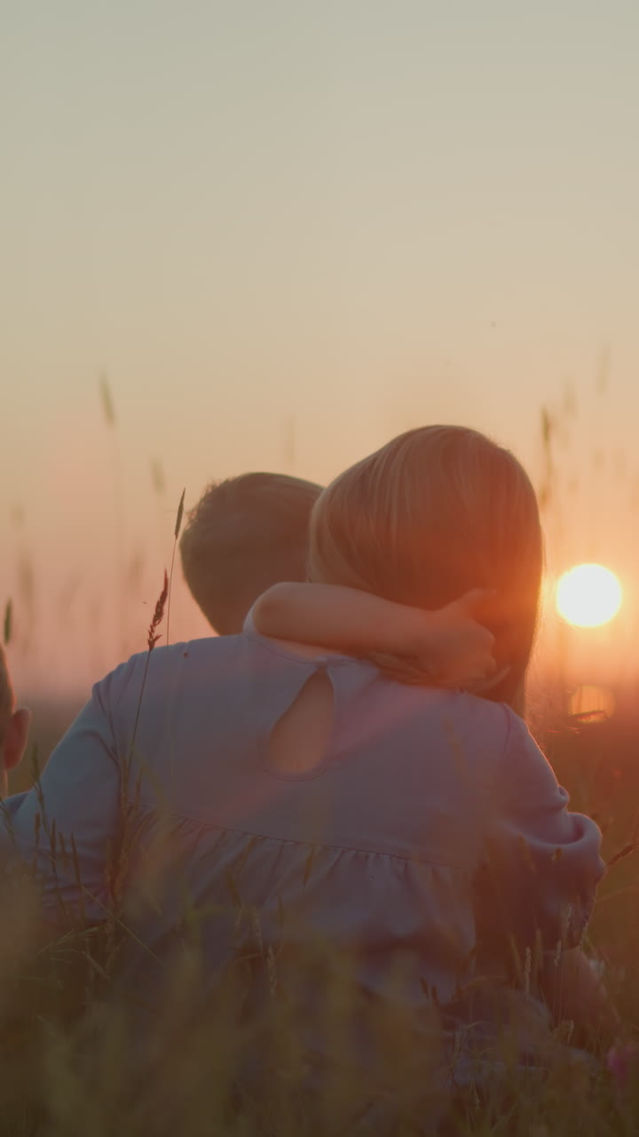 un retrato de una madre con un vestido azul sentada en un campo durante la puesta del sol, sosteniendo a su hijo menor en su regazo mientras el niño mayor se sienta a su lado. todos miran hacia el lago sereno