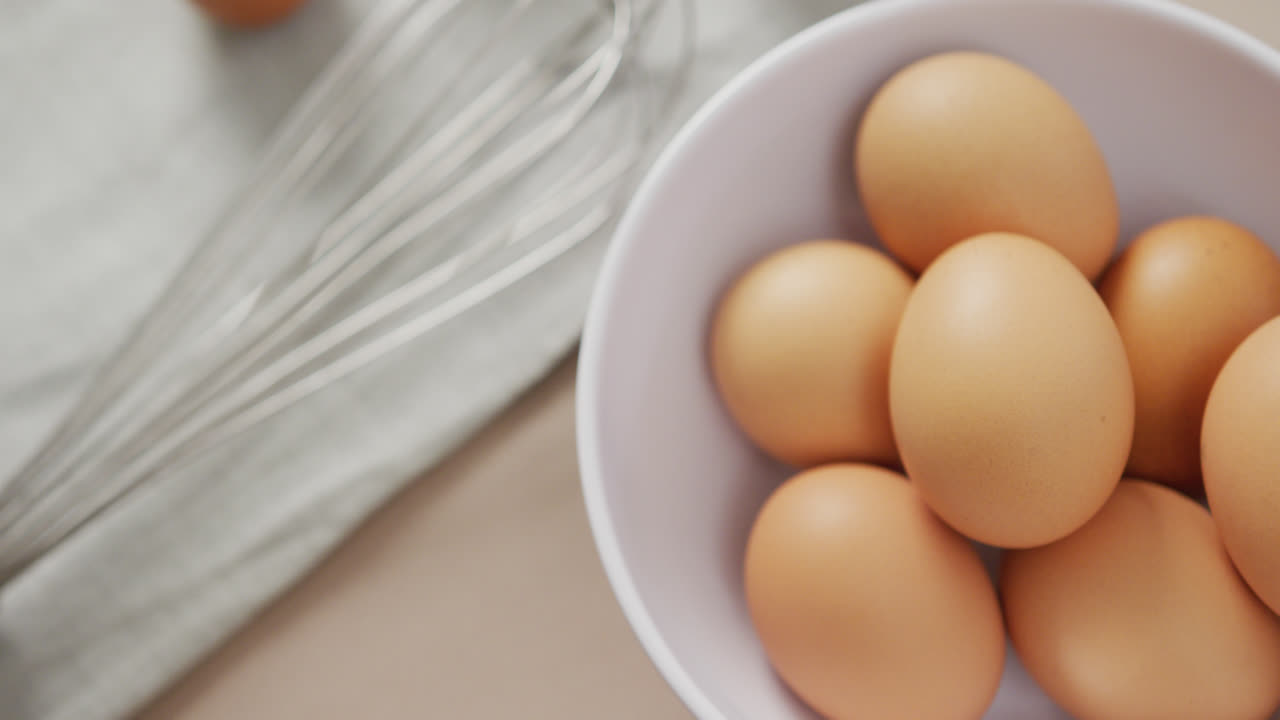Video of overhead view of white bowl with eggs and egg whisk on beige background
