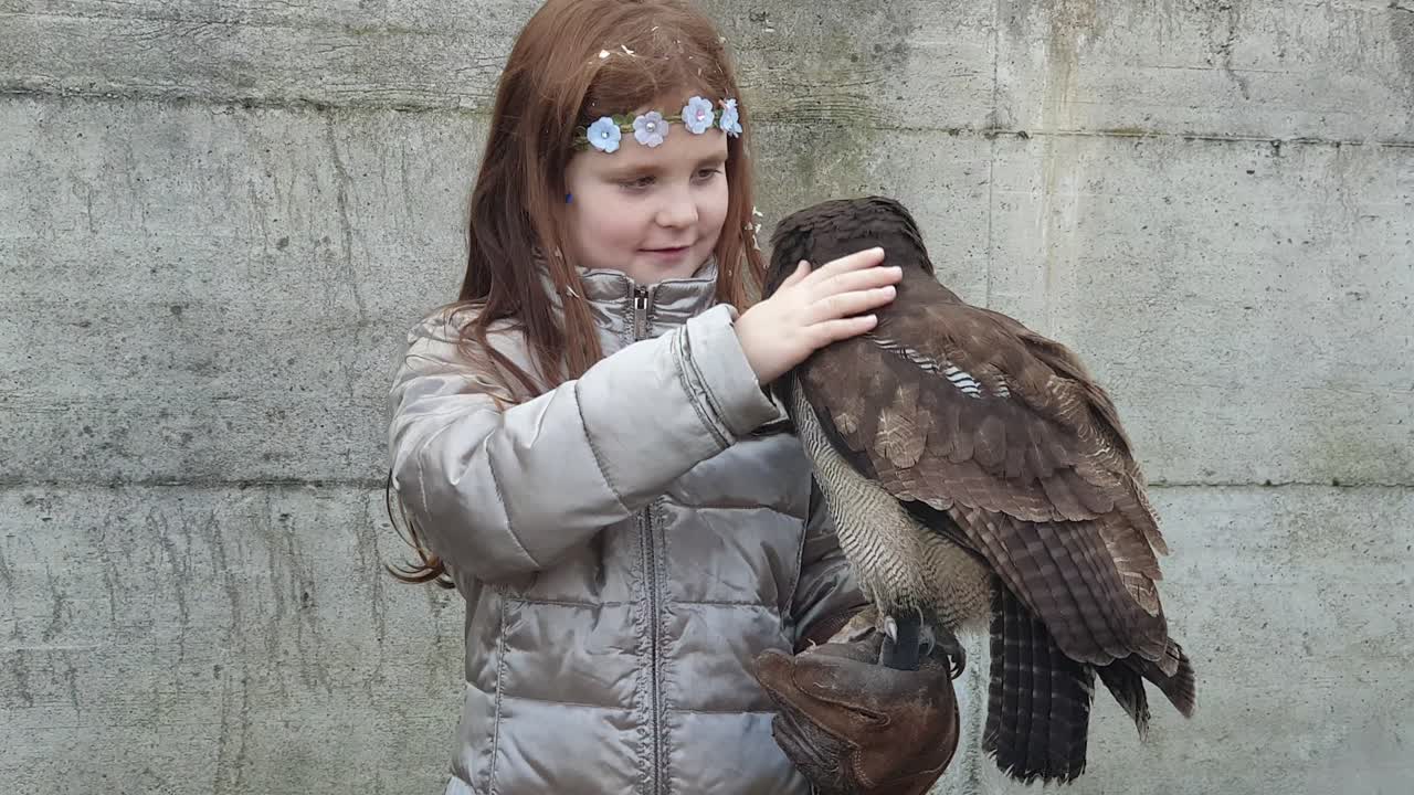 Smiling little girl with crown of flowers caress tawny owl on arm