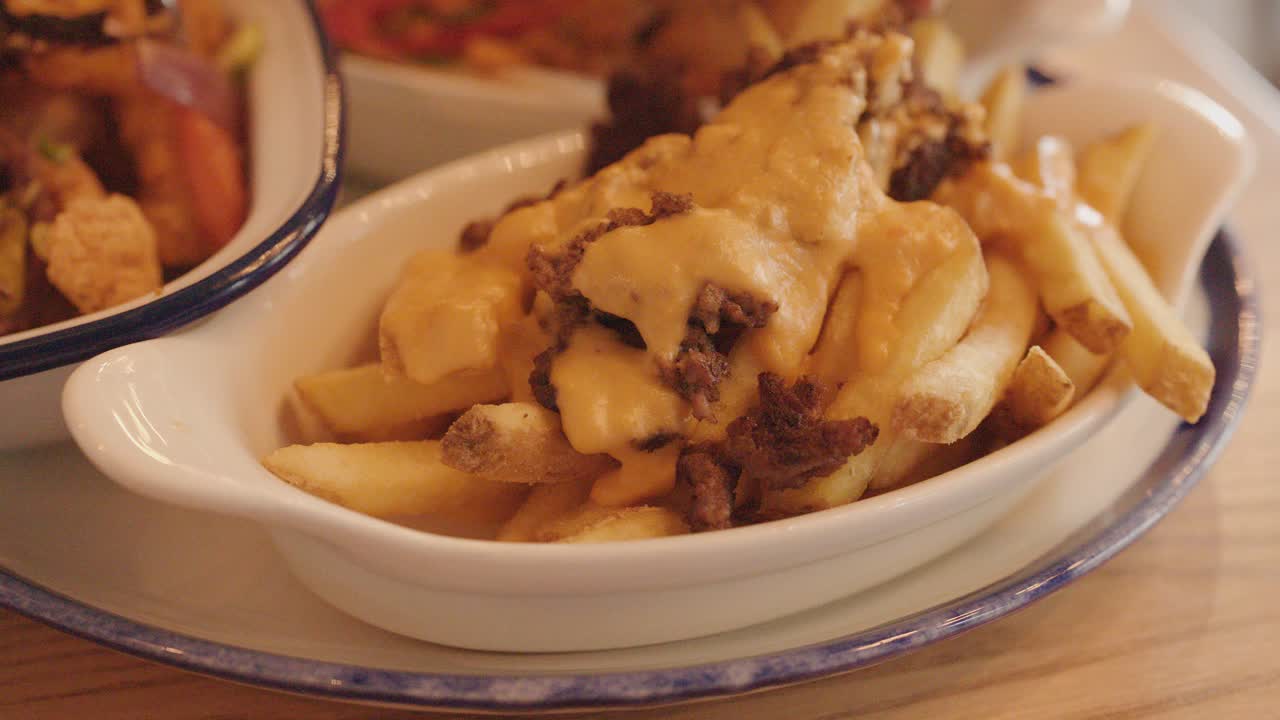 A hand reaches for a dish of loaded fries topped with melted cheese and ground meat, under warm indoor lighting on a wooden table