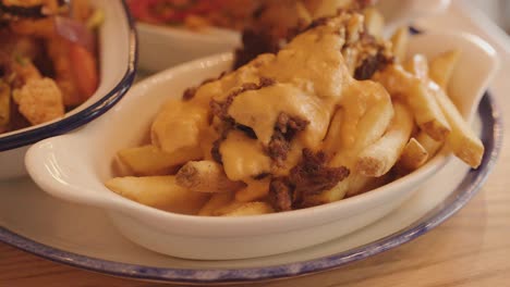 a hand reaches for a dish of loaded fries topped with melted cheese and ground meat, under warm indoor lighting on a wooden table