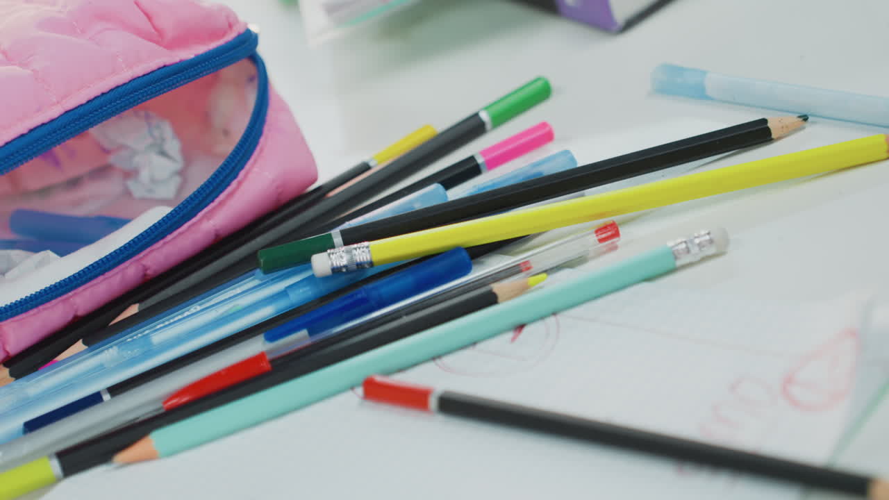 Dağınık kalemler ve uçlarla dolu pembe kalem kutusu, not defteri sayfasının yakın çekim görüntüsü, pastel silgi kırpıntıları ve renkli markörler, grafit uçlar ve kalemtıraş görülebiliyor, dağınık öğrenci çalışma hazırlığı atmosferi