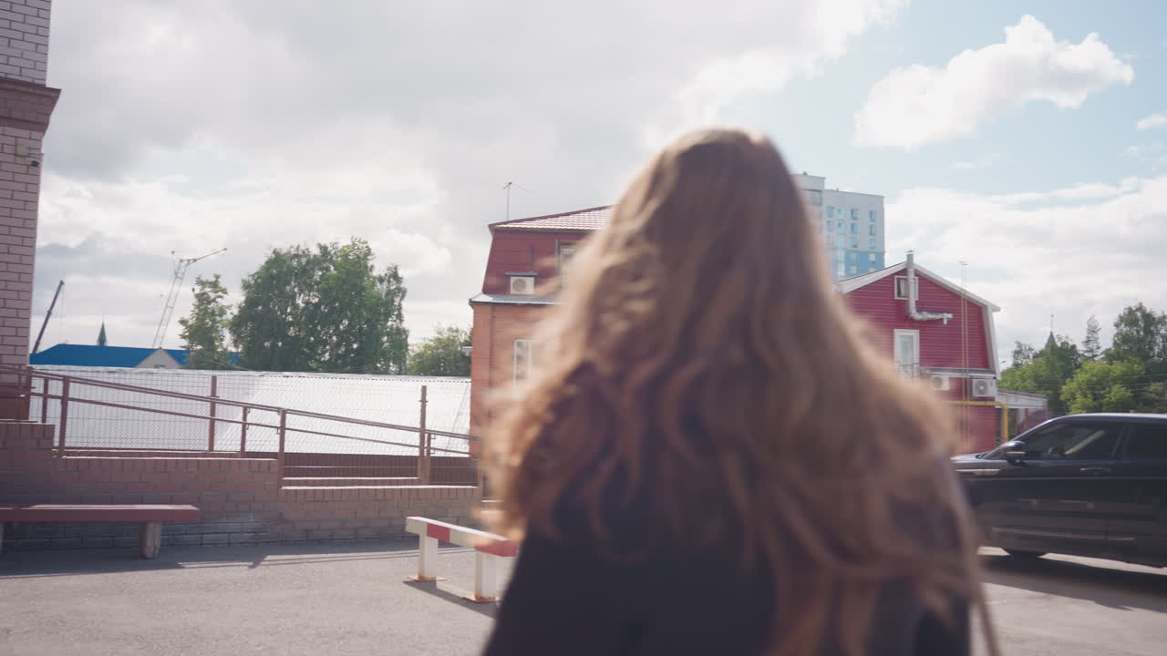 금발 머리 소녀가 갈색 숄더백을 흔들며 벽돌 건물과 난간을 지나 카메라에서 멀어지고 있어요. 아침 햇살이 구름 사이로 비치고, 안뜰과 붉은 지붕의 집이 도시적인 분위기를 연출해요.