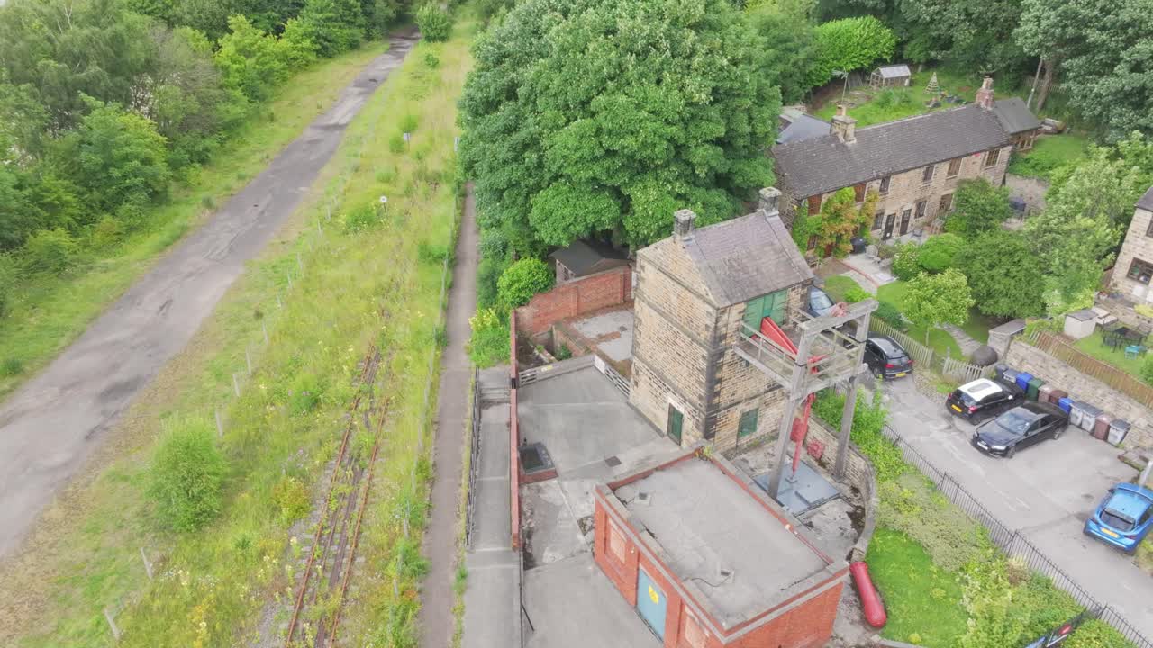 Ascending drone orbit over Elsecar Heritage Centre showing stone and brick workshops, sawtooth roofs, the New Yard chimney, central market tents, parked vehicles and an overgrown disused railway
