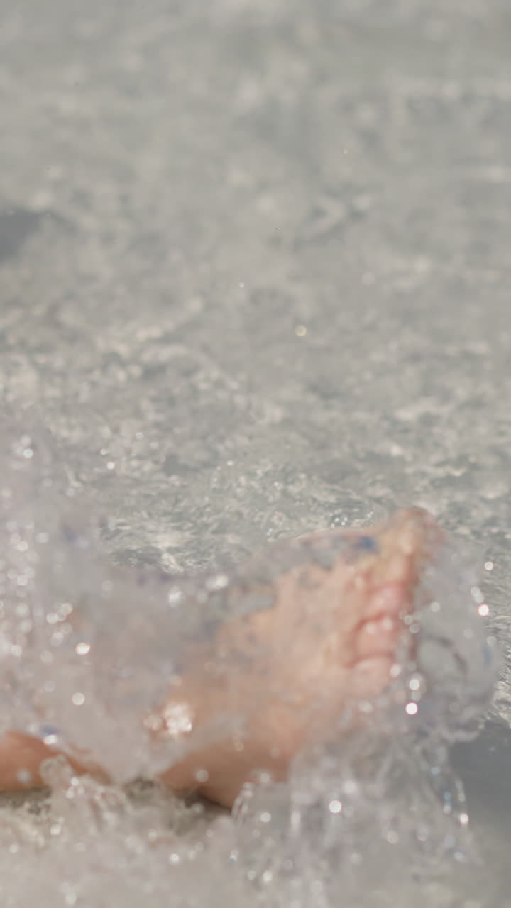 Joyful little child swings legs and splashes clear water in surfline on wet shore at tropical resort on summer day close view slow motion