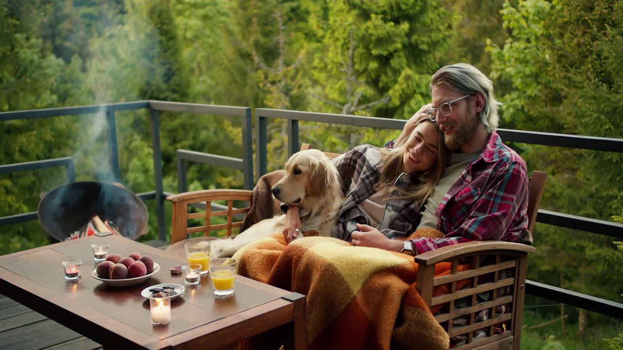 A romantic evening in a country house: a blond man and a blond girl are sitting on a couch wrapped in a blanket. Near them sits their light-colored dog. Relax with a view of the mountains and the forest