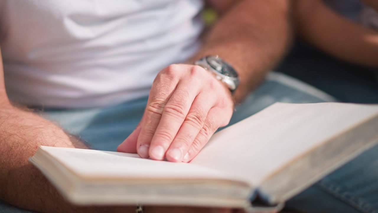 Close-up de homem lendo livro aberto com a mão apoiada na página, vestindo roupas casuais e relógio. A cena mostra iluminação suave destacando detalhes como a textura da pele em um ambiente natural.