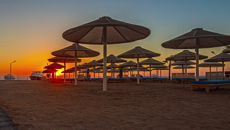 Empty-hotel-lounge-chairs-and-tiki-umbrellas-on-beach-as-sunset-glows-in-sky,-Timelapse