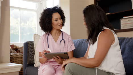 nurse in pink scrubs showing tablet to woman sitting on couch, discussing health