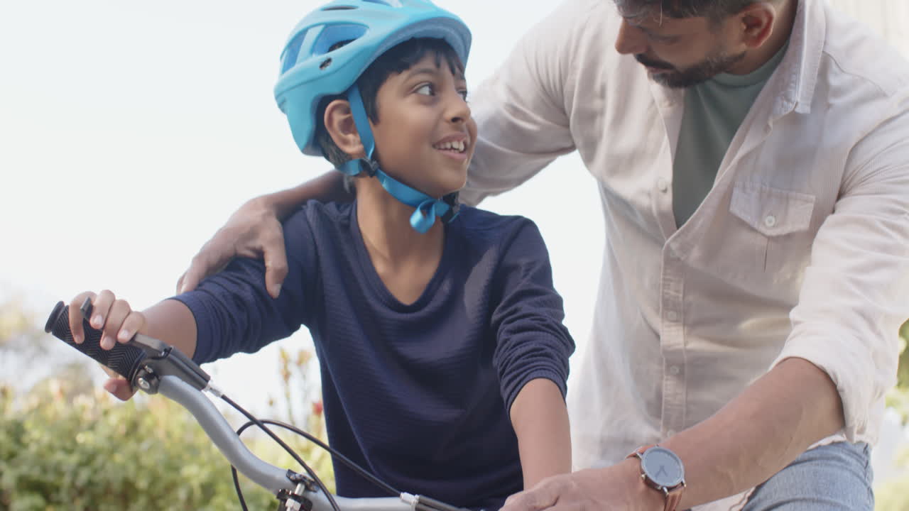 Indian father teaching young son to ride bicycle, bonding outdoors with helmet on