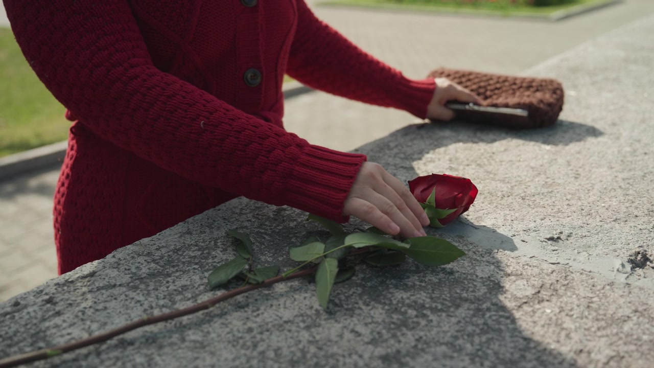 Una donna con un maglione rosso si avvicina, posa la sua pochette marrone su un davanzale di pietra e tocca o sistema delicatamente una rosa fresca con foglie verdi. La scena presenta una luce naturale soffusa che evidenzia i dettagli.