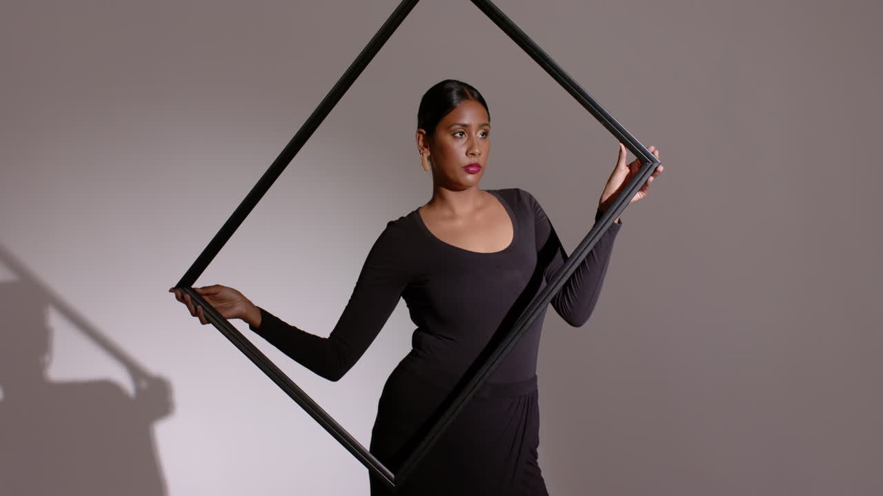 Lifting black frame from waist, Indian woman rotating frame, aligning face against gray studio wall