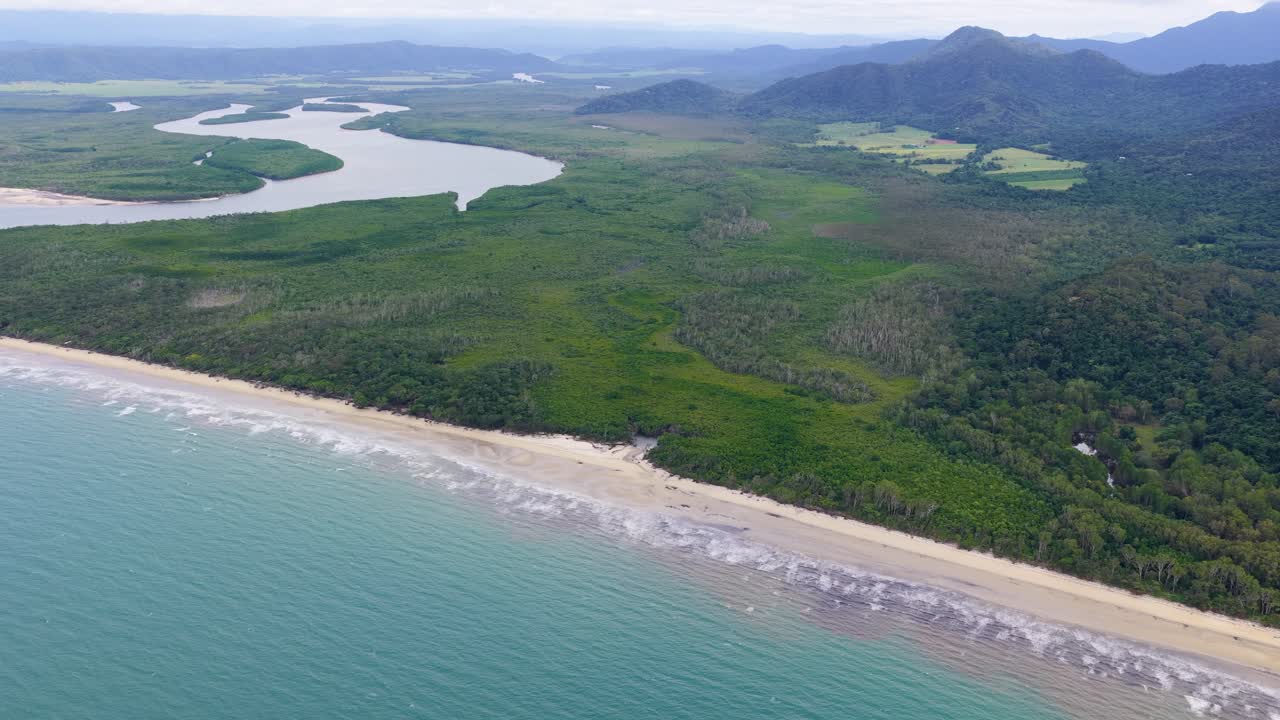 Drone footage glides above the lush Daintree Rainforest coastline, revealing winding rivers, dense jungle, sandy beaches, and distant mountains under soft daylight
