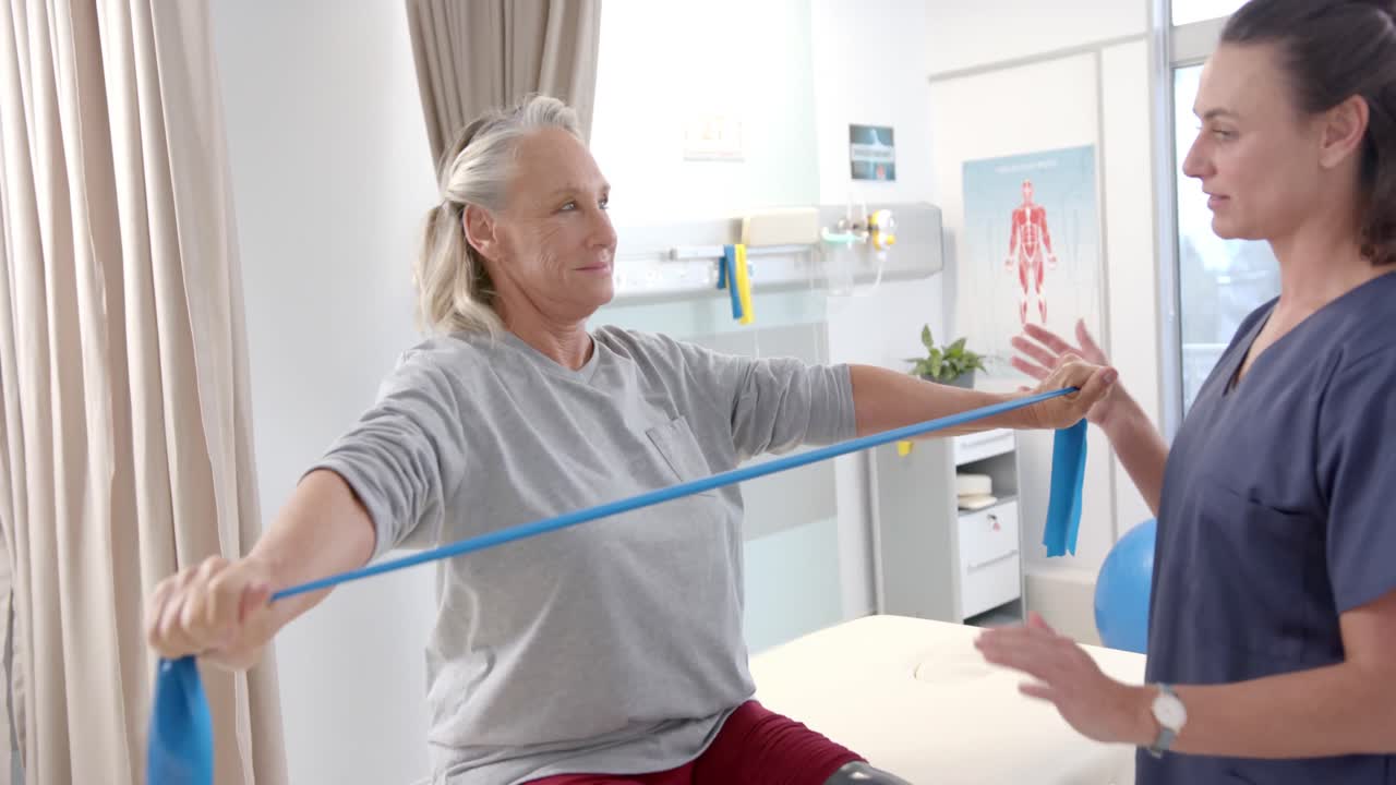 백인 여성 물리 치료사가 여성 노인 환자에게 저항 밴드로 운동을 도와줍니다.