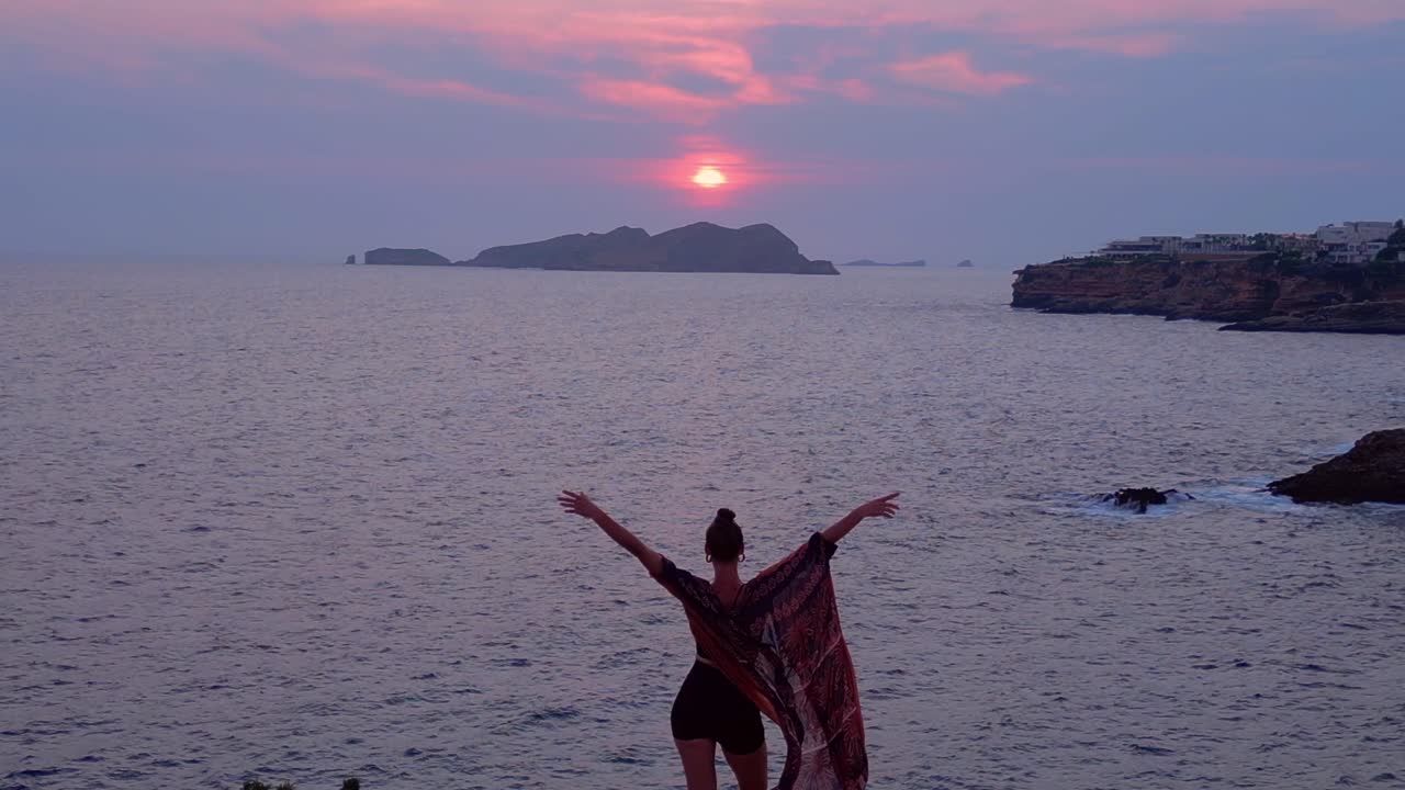Woman performing heart gesture with hands capturing setting sun over ocean, promoting wellness and mindfulness. Best aerial view flight descending drone