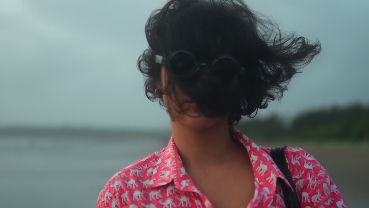 Primo piano di donna in spiaggia, con i capelli mossi dal vento sul viso, che indossa occhiali da sole