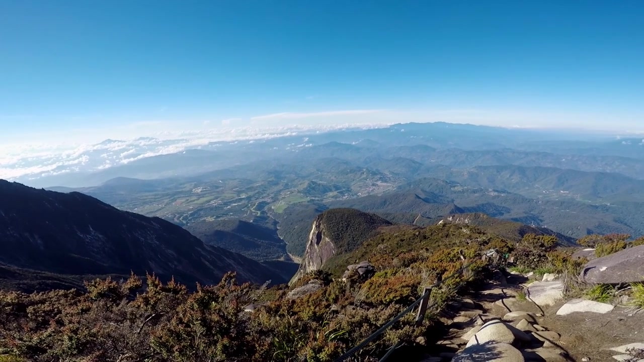 Amplia toma panorámica descendente desde el sendero escarpado y la vegetación alpina del Monte Kinabalu sobre el extenso valle de Kundasang y las cordilleras distantes bajo la luz brillante del sol.
