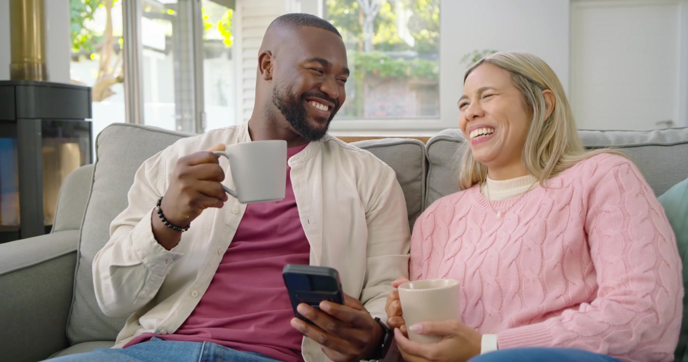 Coppia afroamericana sul divano, l'uomo che guarda il telefono mostrando lo schermo, che legano, lei che sorseggia da una tazza e ride.