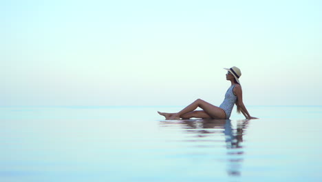 Stilvolle-Frau-In-Badeanzug-Und-Hut-Am-überlaufpool-Am-Pool-Mit-Himmlischer-Skyline-Im-Blick