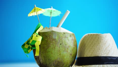 straw hat and a fresh young coconut coconut tropical drink with mini umbrella and ribbon ketupat against blue background