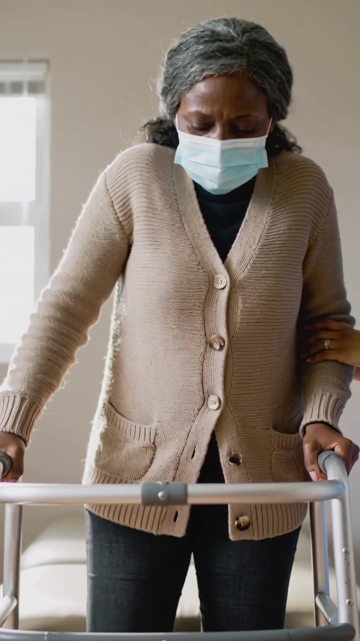 Vertikal video: Eldre person med munnbind griper rullator, reiser seg fra sete, mens sykepleier holder armen.