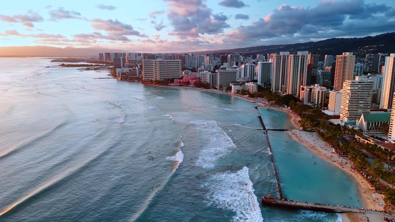 As ondas do Oceano Pacífico rolam para a praia de areia de Waikiki, Honolulu, Havaí, EUA. Vista aérea da cidade ao pôr do sol. Céu nublado ao fundo