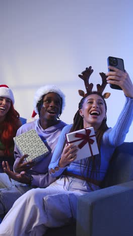 Vertikale-Videostudioaufnahme-Von-Freunden-Der-Generation-Z-Zu-Weihnachten,-Die-Mit-Weihnachtsmütze-Und-Rentiergeweih-Auf-Dem-Sofa-Sitzen-Und-Ein-Selfie-Mit-Dem-Mobiltelefon-Machen-2