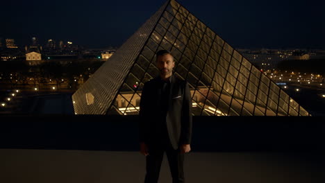 man in suit posing in front of the louvre pyramid at night