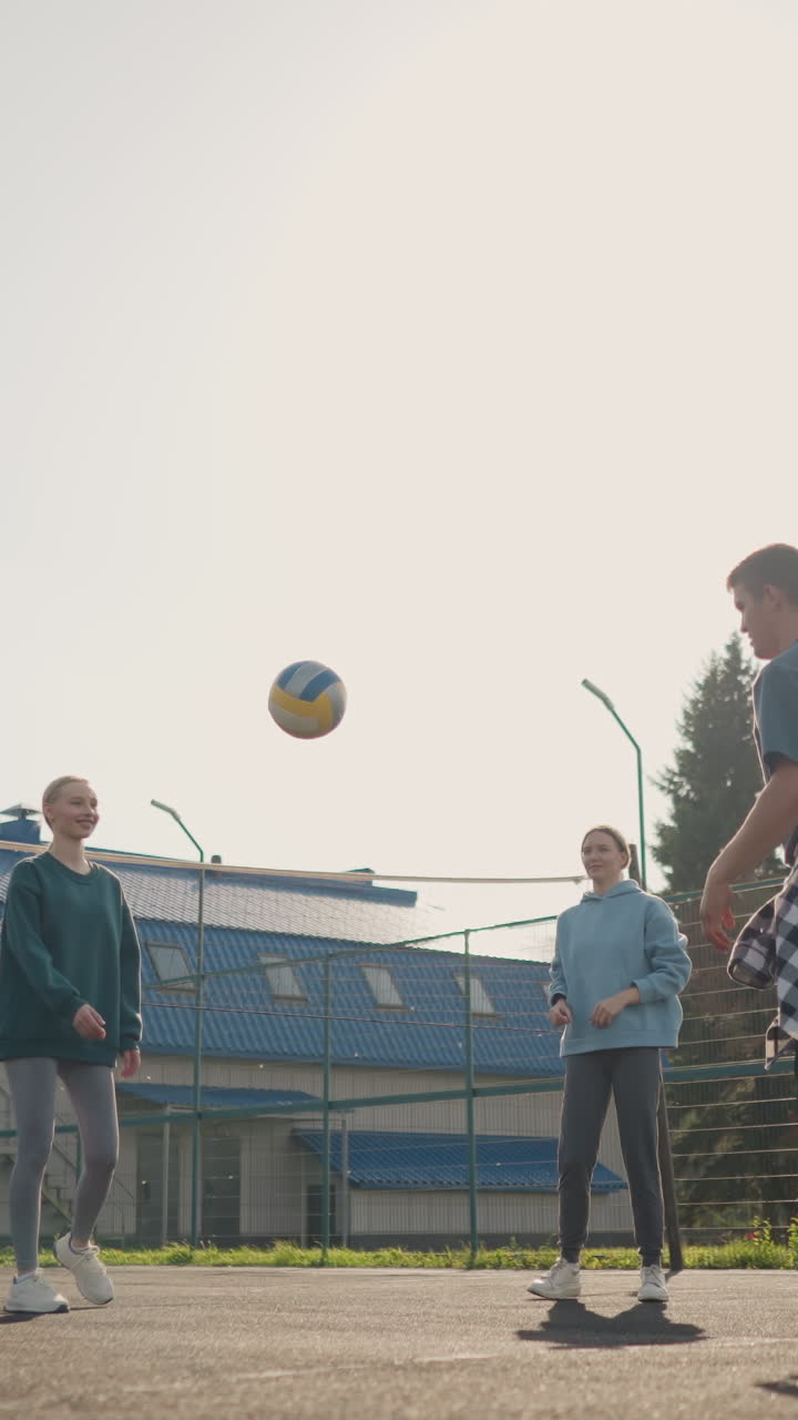 코치가 야외 코트에서 배구하는 세 명의 여자 선수와 함께 훈련 세션을 이끌고, 코치가 건물과 울타리를 배경으로 가져오기 위해 손을 는 동안 공이 땅에 떨어집니다.